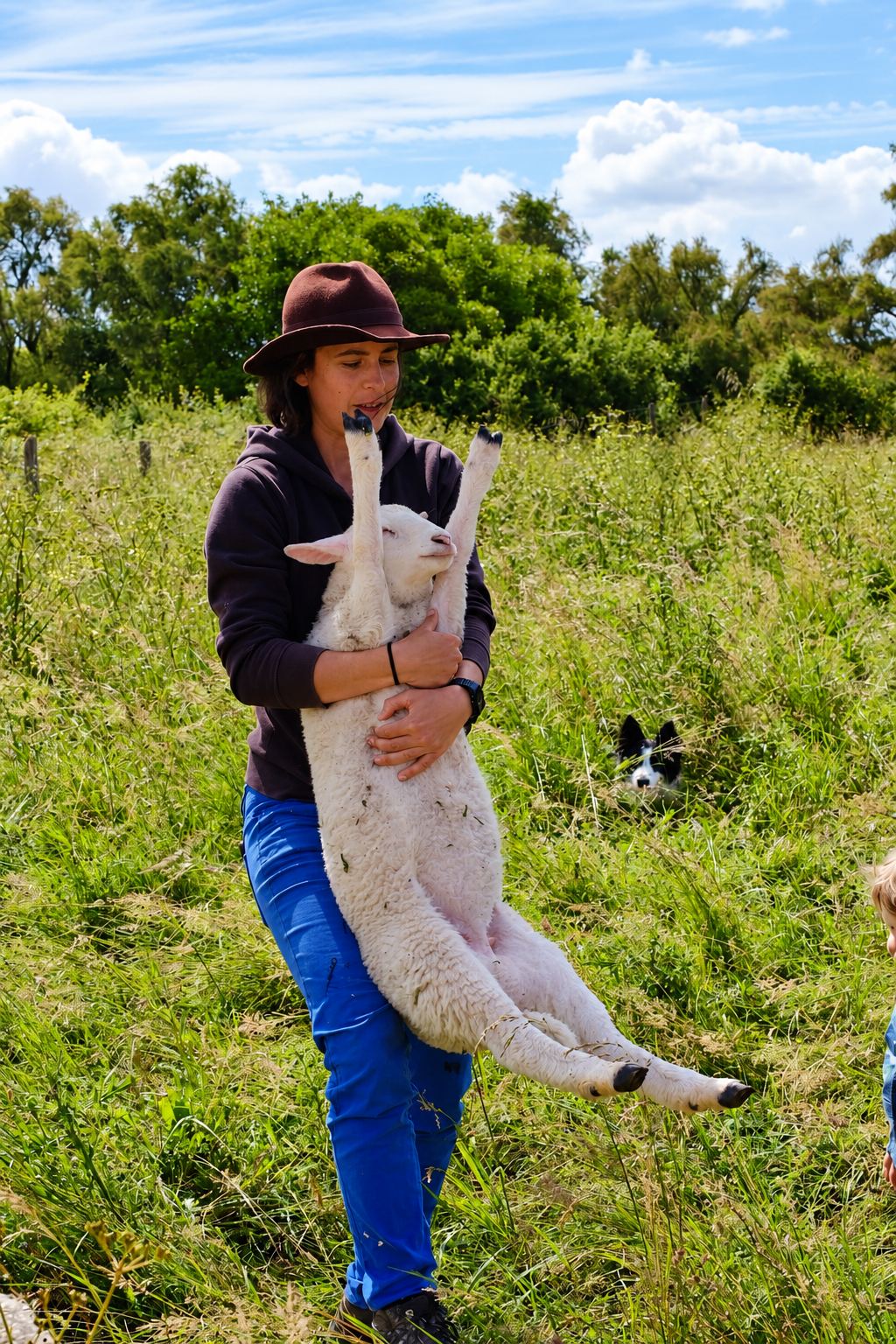 Une femme coiffée d'un chapeau marron porte un mouton blanc dans ses bras au milieu d'un pré herbeux, avec un chien noir et blanc caché dans l'herbe derrière elle.