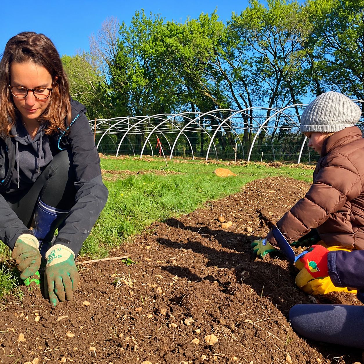 Une femme portant des gants de jardinage et des enfants travaillent la terre d'une planche de culture, avec une serre tunnel et des arbres en arrière-plan.