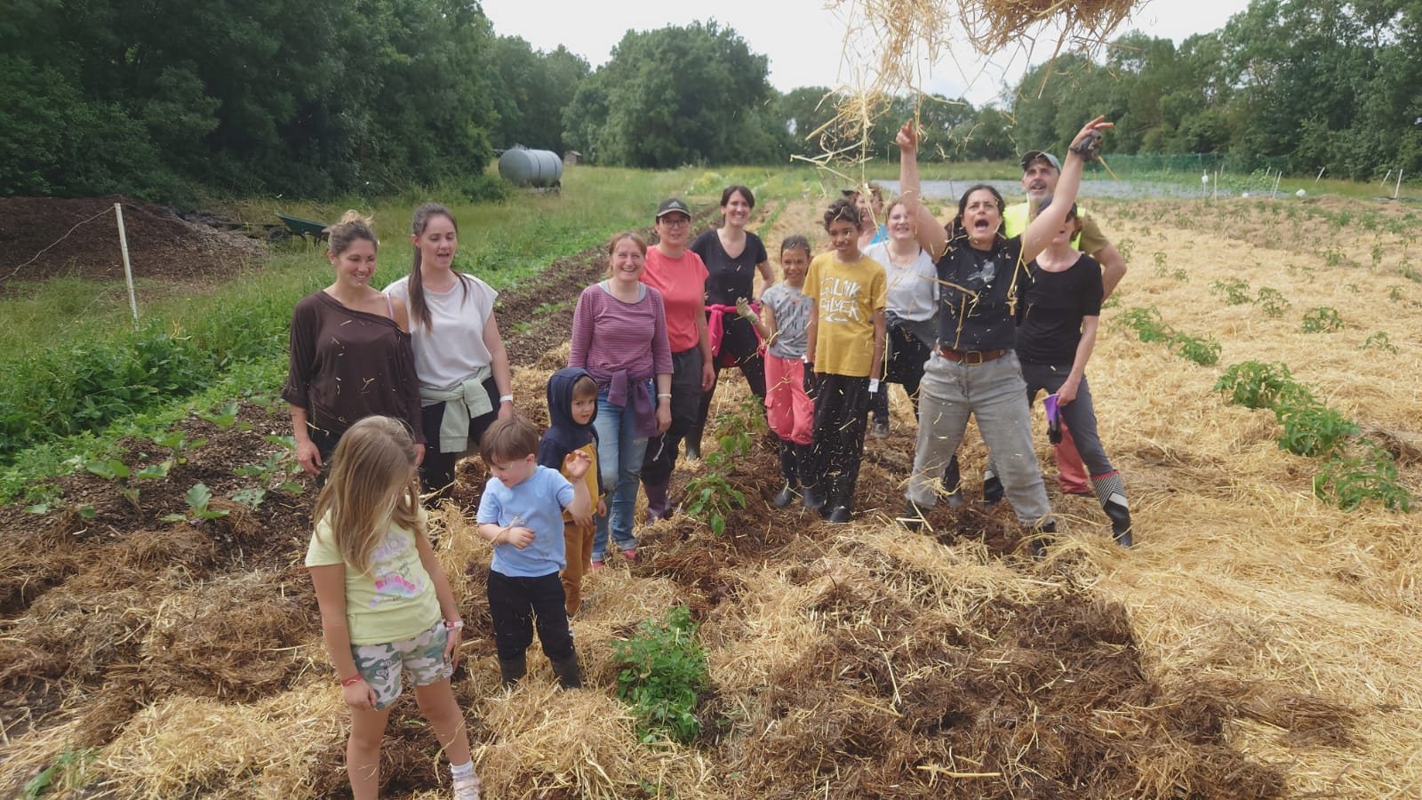 Un groupe d'adultes et d'enfants souriants pose dans un champ de cultures recouvert de paille, une femme lançant de la paille en l'air.
