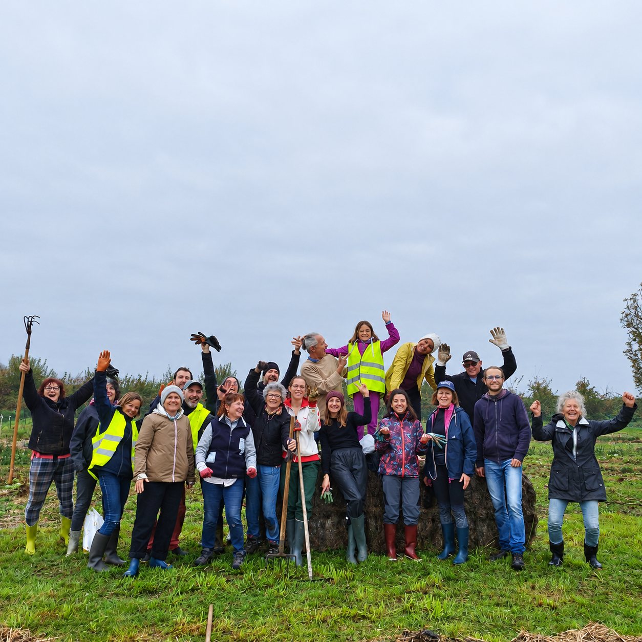 Un groupe de personnes en tenue de travail, certaines avec des gilets jaunes et des outils de jardinage, lèvent les bras en riant dans un champ sous un ciel brumeux.