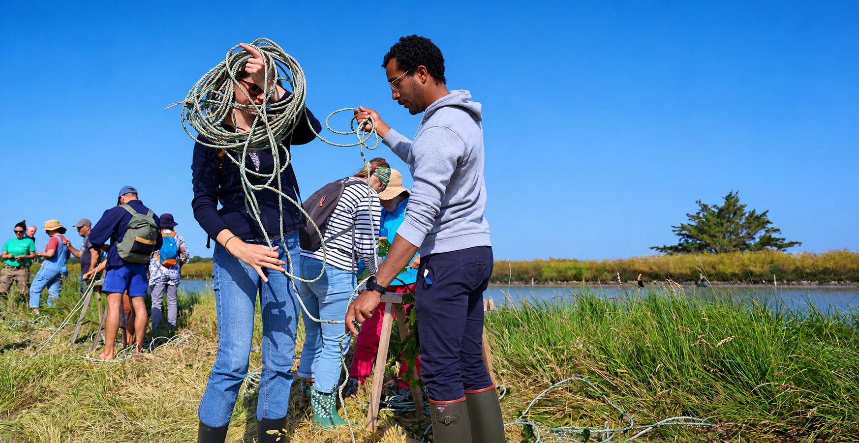 Une femme et un homme démêlent une longue corde au bord d'un marais, entourés d'un groupe de personnes sous un ciel bleu.