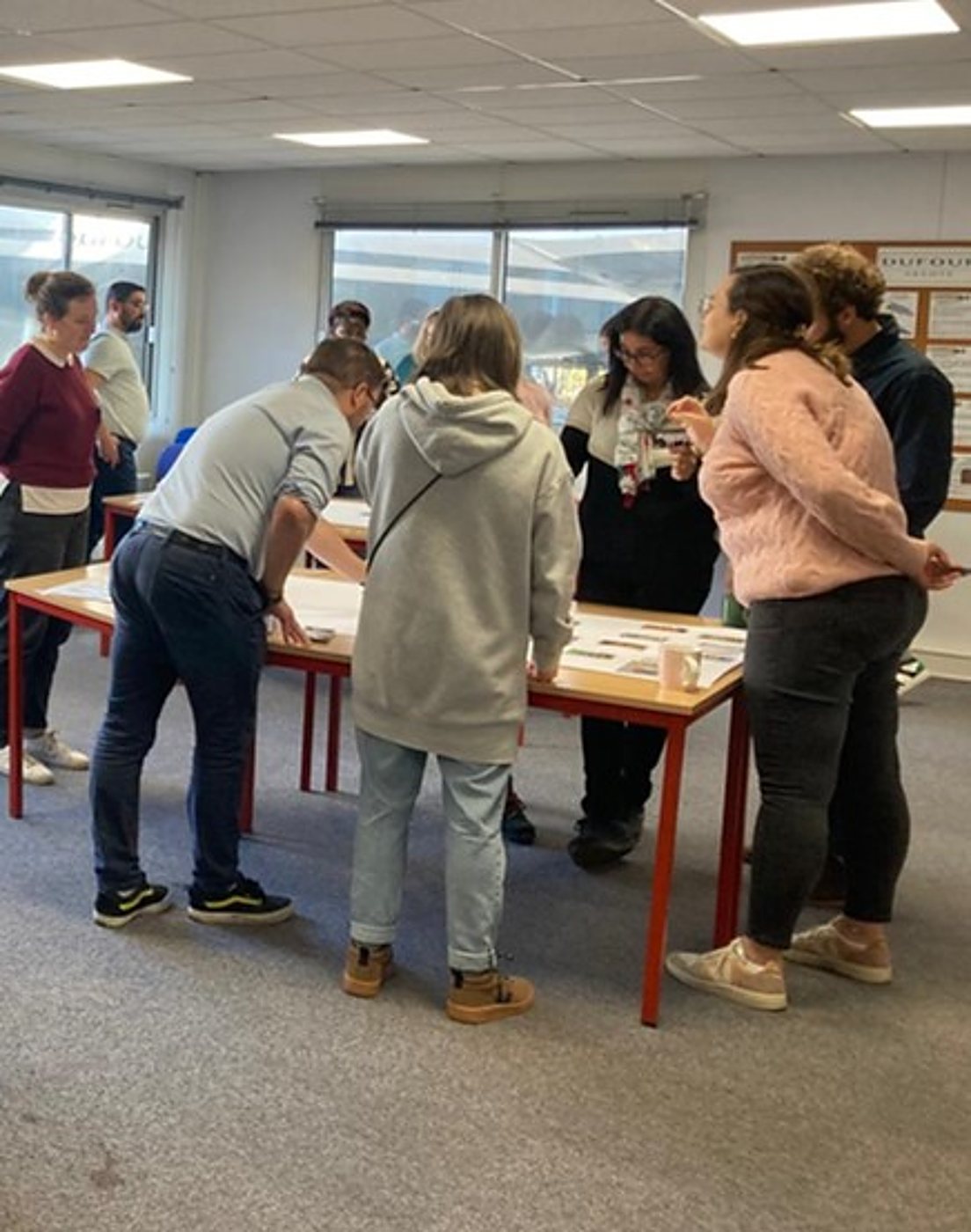 Plusieurs personnes debout autour d'une grande table aux pieds rouges, examinant des documents dans une salle claire aux grandes fenêtres.