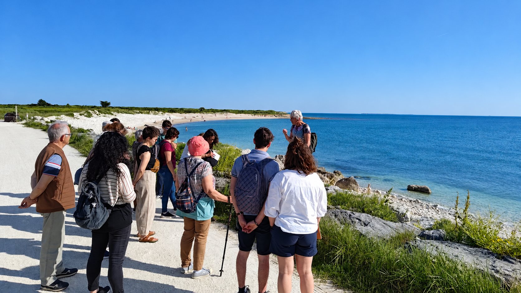 Un groupe de personnes écoute une guide en randonnée sur un sentier au bord de la mer, près d'une plage sous un ciel bleu.