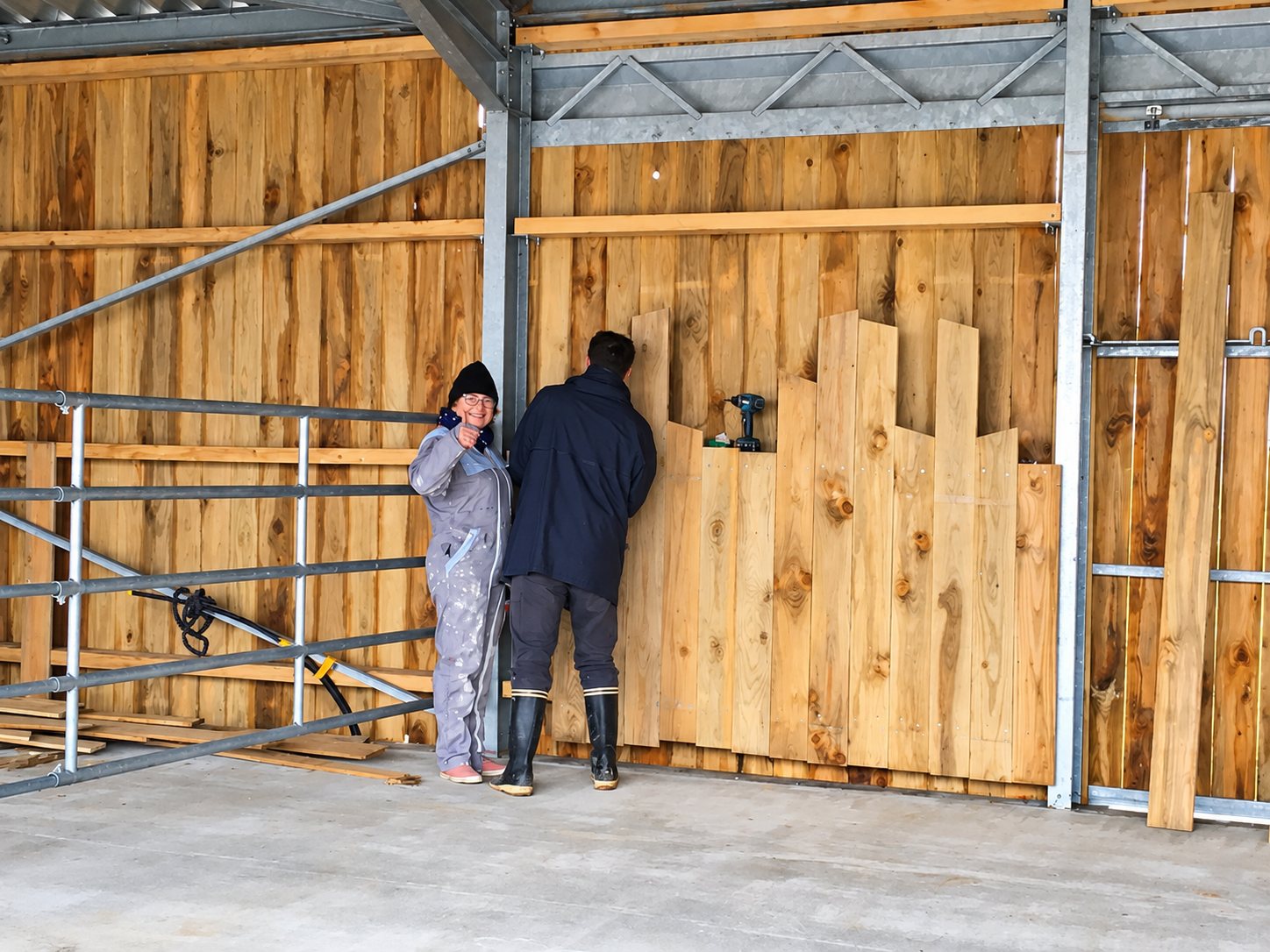 Deux personnes installent des planches de bois brut sur la paroi d'un bâtiment à ossature métallique, une perceuse posée sur les planches.