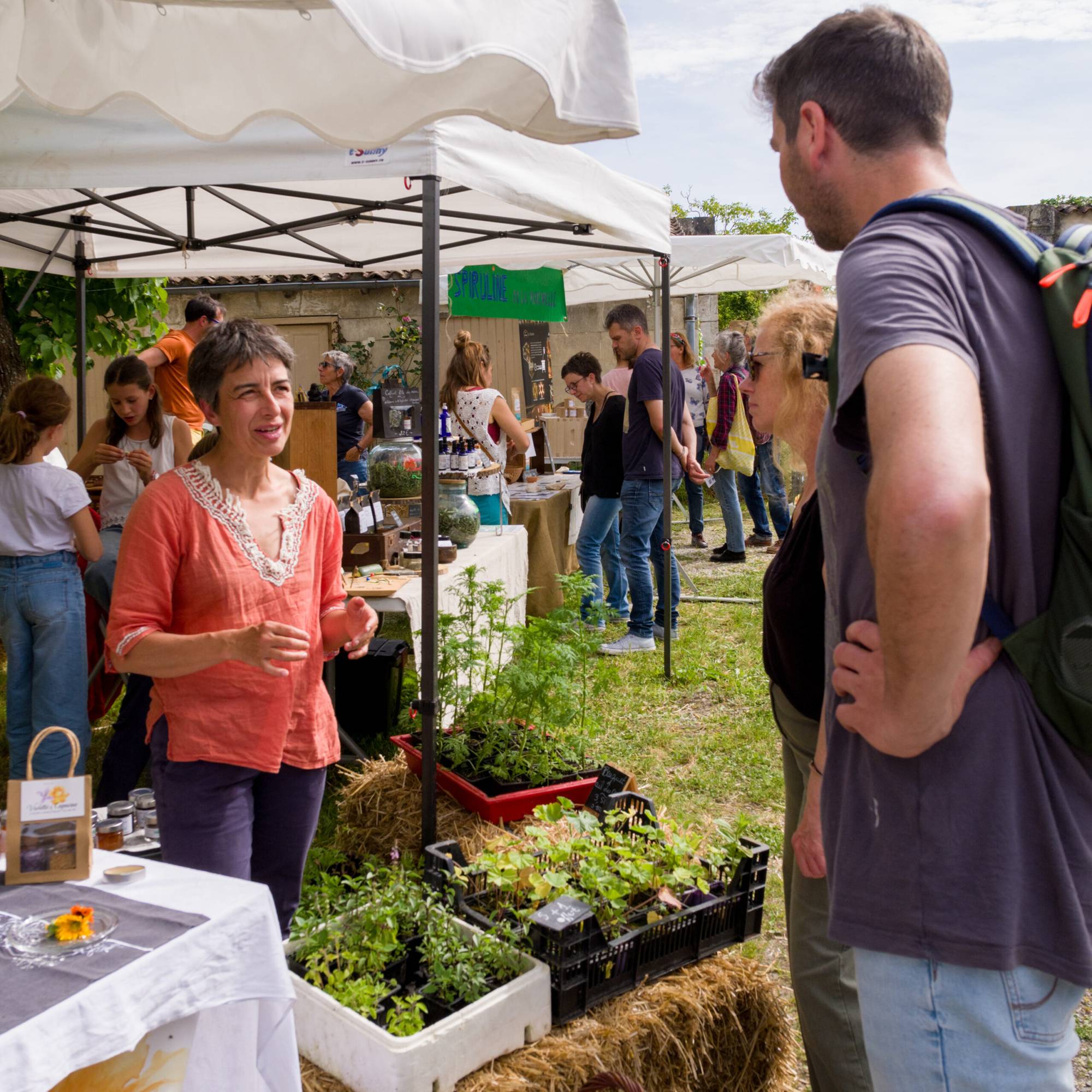 Une femme en blouse corail parle à des visiteurs devant des tables de plants aromatiques et de pots de miel sous des tentes blanches lors d'un marché en plein air.